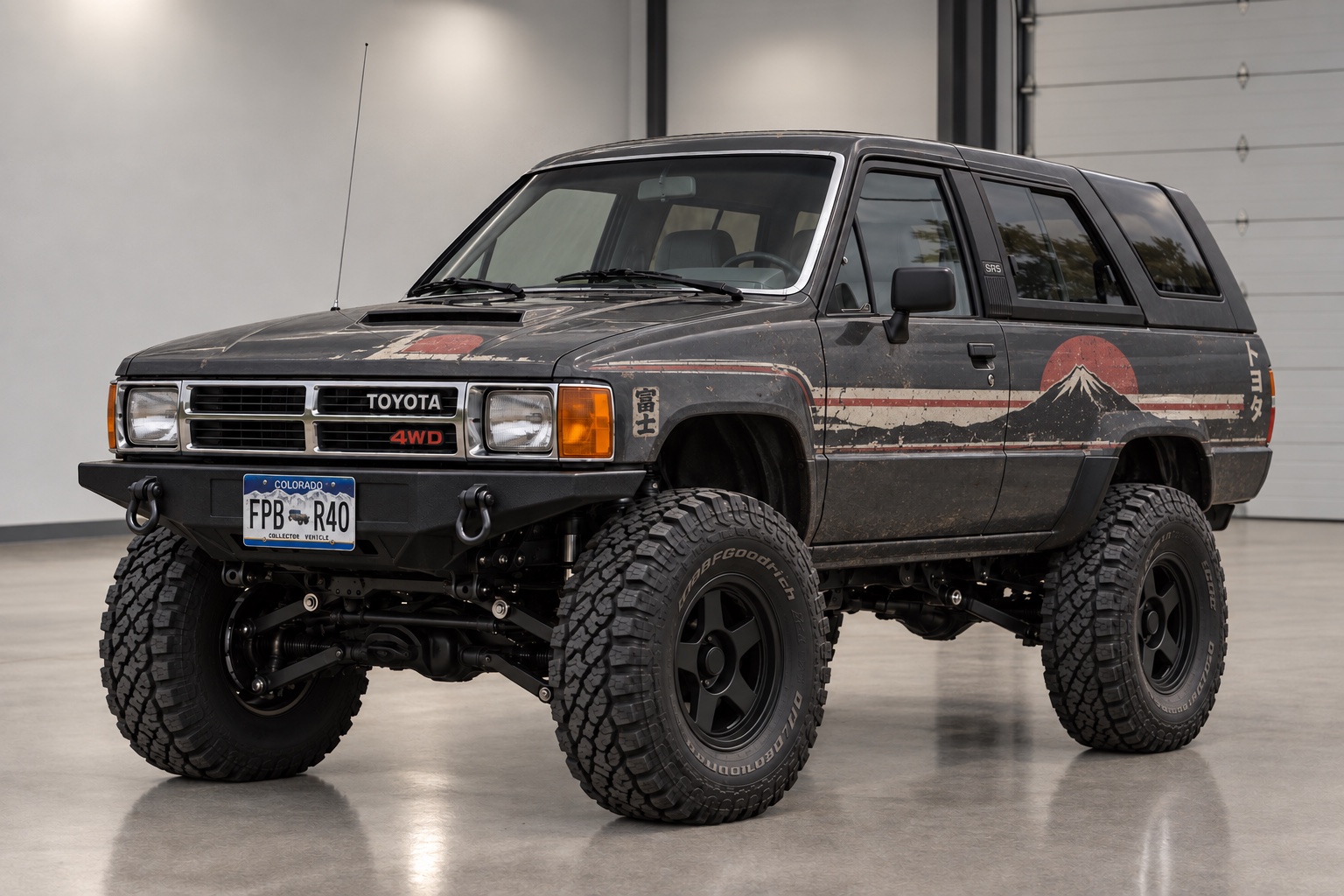 The 4Runner front three quarter in a workshop, matte black with the weathered Mount Fuji and rising sun livery down the side, on a lift and BFGoodrich all terrains