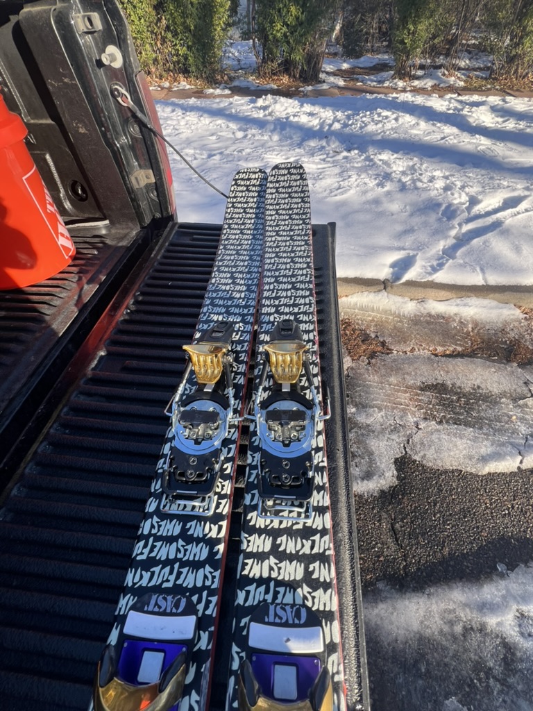 A pair of skis with painted topsheets and bindings fitted, propped in a truck bed against a mountain sky