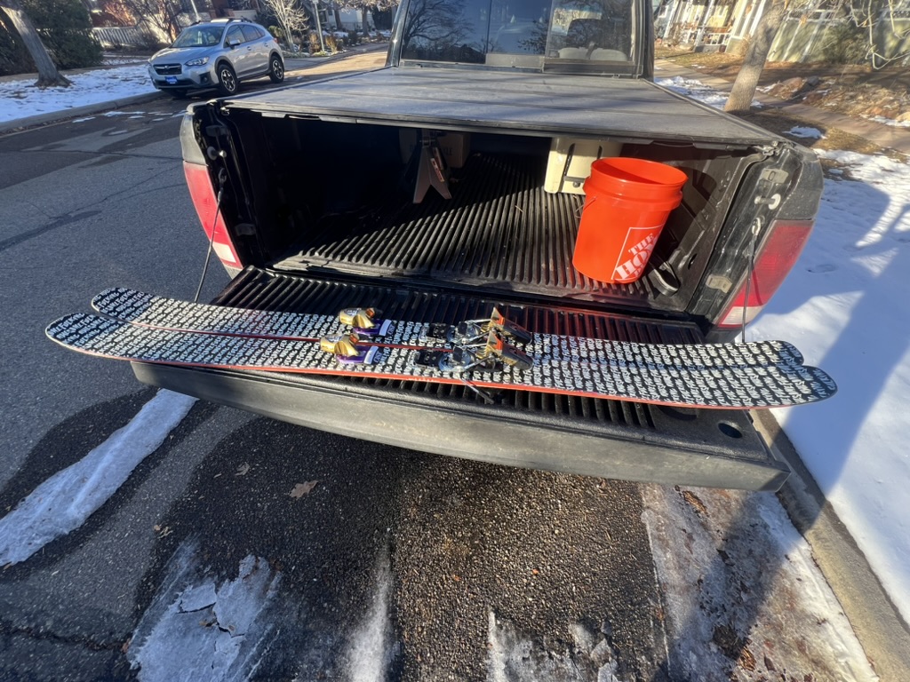 Skis with repeating hand lettered graphics laid across an open tailgate, an orange bucket in the truck bed behind them